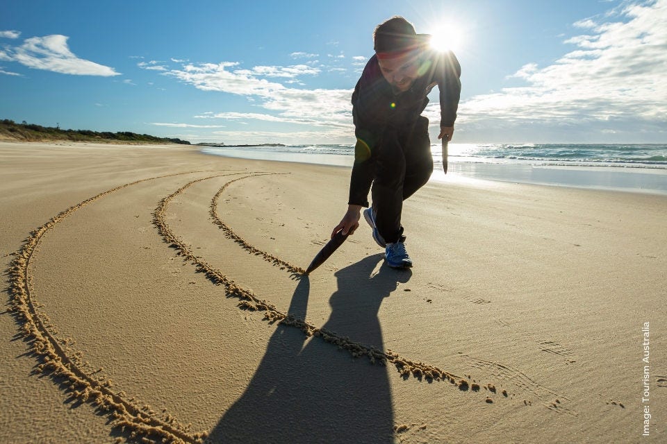 An Indigenous guide from Ngaran Ngaran Cultural Awareness draws in the sand of Callala Bay Shoalhaven with a wooden knife. Image: Tourism Australia.